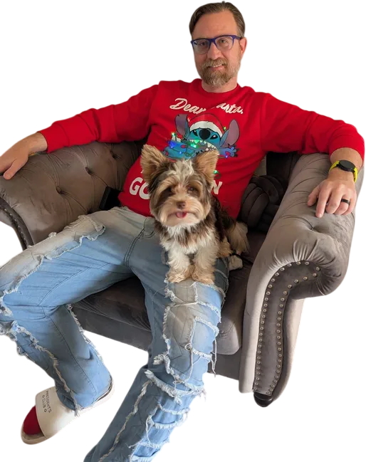 Man wearing a red cartoon Christmas sweater and ripped jeans sits relaxed on a gray armchair with a small dog on his lap.